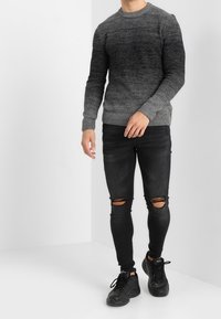 Grauer, Verlauf gestrickter Pullover mit rundem Hals, kombiniert mit schwarzen, abgerissenen Skinny-Jeans und schwarzen Sport Sneakers, vor einem weißen Hintergrund.