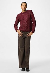 Burgundijas adīta džemperis ar kabeļu rakstu, ar apaļu kaklu un garām piedurknēm, savienots ar plašām brūnām biksēm un melniem apaviem.