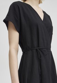 Robe portefeuille noire en tissu léger. Dotée de manches courtes, d'un décolleté en V profond et d'une ceinture en tissu à la taille. Texture lisse.