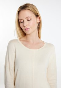 Beige Strickpullover mit weitem Rundhalsausschnitt und leicht herunterhängenden Schultern, der eine glatte Textur und ein zentrales Nahtdetail aufweist.