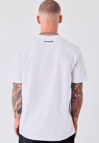 Hombre con cabello corto y brazos tatuados, vistiendo una camiseta blanca de PXP, collar de cadena plateada y aretes, de espaldas a la cámara.