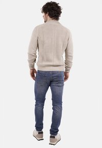 Uomo con capelli ricci che indossa un maglione beige a coste, jeans blu e sneakers bianche, in piedi di spalle su uno sfondo bianco.