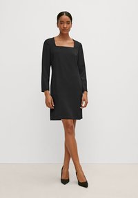 Schwarzes Kleid mit quadratischem Ausschnitt, langen Ärmeln und geradem Schnitt. Glattes Material und seitliche Schlitze. Getragen mit spitzen schwarzen Pumps.