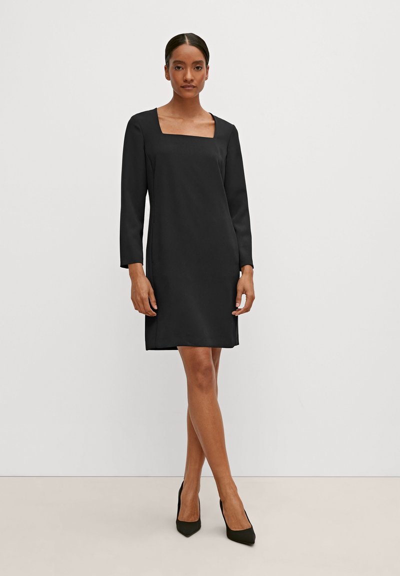 Schwarzes Kleid mit quadratischem Ausschnitt, langen Ärmeln und geradem Schnitt. Glattes Material und seitliche Schlitze. Getragen mit spitzen schwarzen Pumps.