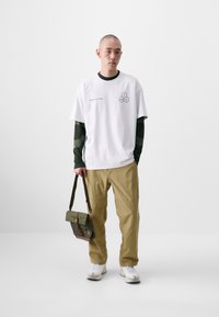 White T-shirt with graphic and text, long green-striped sleeves underneath, tan pants, white sneakers, and an olive green messenger bag.