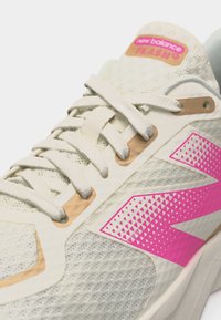 Αθλητικό παπούτσι New Balance Flash σε λευκό και μπεζ με ροζ λογότυπο "N" και ροζ ετικέτα στη γλώσσα, κοντινή λήψη στα κορδόνια και το πλεκτό υλικό.