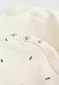 Vaaleanbeige puuvillahaalari, jossa on mustia pistekuvioita. siinä on pyöreä pääntie ja kolme hopeista nepparia selässä, mikä helpottaa pukemista.