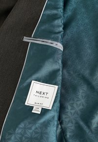 Intérieur d'un blazer ajusté avec une doublure à motifs turquoise, des accents gris et une étiquette avec "NEXT TAILORING" et les détails de taille.