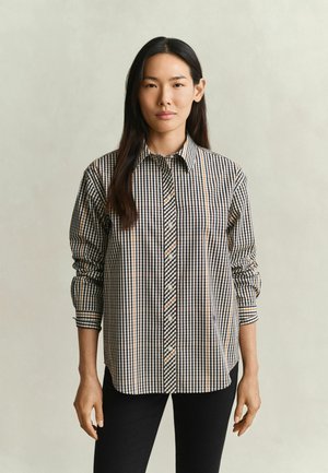 Femme aux longs cheveux foncés portant une chemise à carreaux noire, blanche et beige à manches longues et un pantalon noir sur un fond uni.