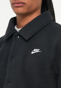 Črna flis jakna z gumbasto zapiranje, ostrim ovratnikom in belim vezenim logotipom Nike na levi strani. Mehak, teksturiran material.