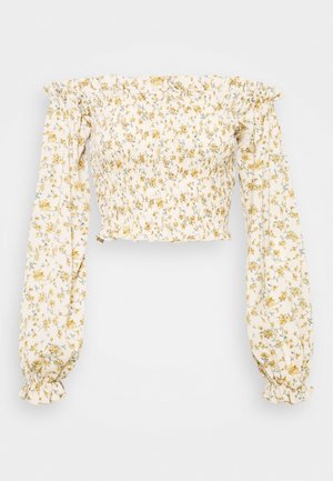 Hvid cropped top med elastisk halsudskæring uden skuldre, lange pufærmer og gult blomsterprint på tekstureret stof.
