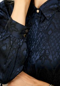 Jūras zila blūze ar fakturētu rakstu, ar apkaklīti un pogu akcentiem uz aproces. Gluda satīna līdzīga materiāla.