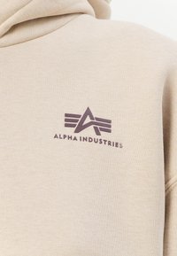 Felpa beige con una texture morbida e un logo frontale che presenta il nome del marchio "ALPHA INDUSTRIES" in viola scuro, affiancato da due strisce orizzontali.