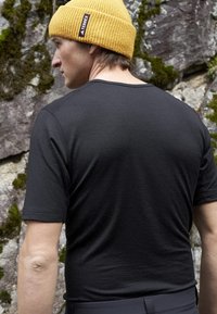 Svart kortärmad skjorta med en jämn textur, med raglanärmar. Bärs tillsammans med en gul ribbad mössa, mot en stenig bakgrund.