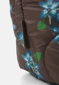 Quiltad brun tyg med ett blommönster i blått och grönt. Inkluderar en brun etikett med text längs kanten.