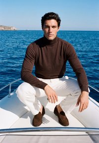 Homme portant un col roulé marron et un pantalon blanc assis sur un bateau avec une mer bleue calme et un ciel clair en arrière-plan.