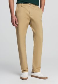 Khaki dopasowane chinosy wykonane z tkaniny bawełnianej. Posiadają klasyczny pięciopocketowy fason, zapięcie na guzik oraz gładką teksturę.