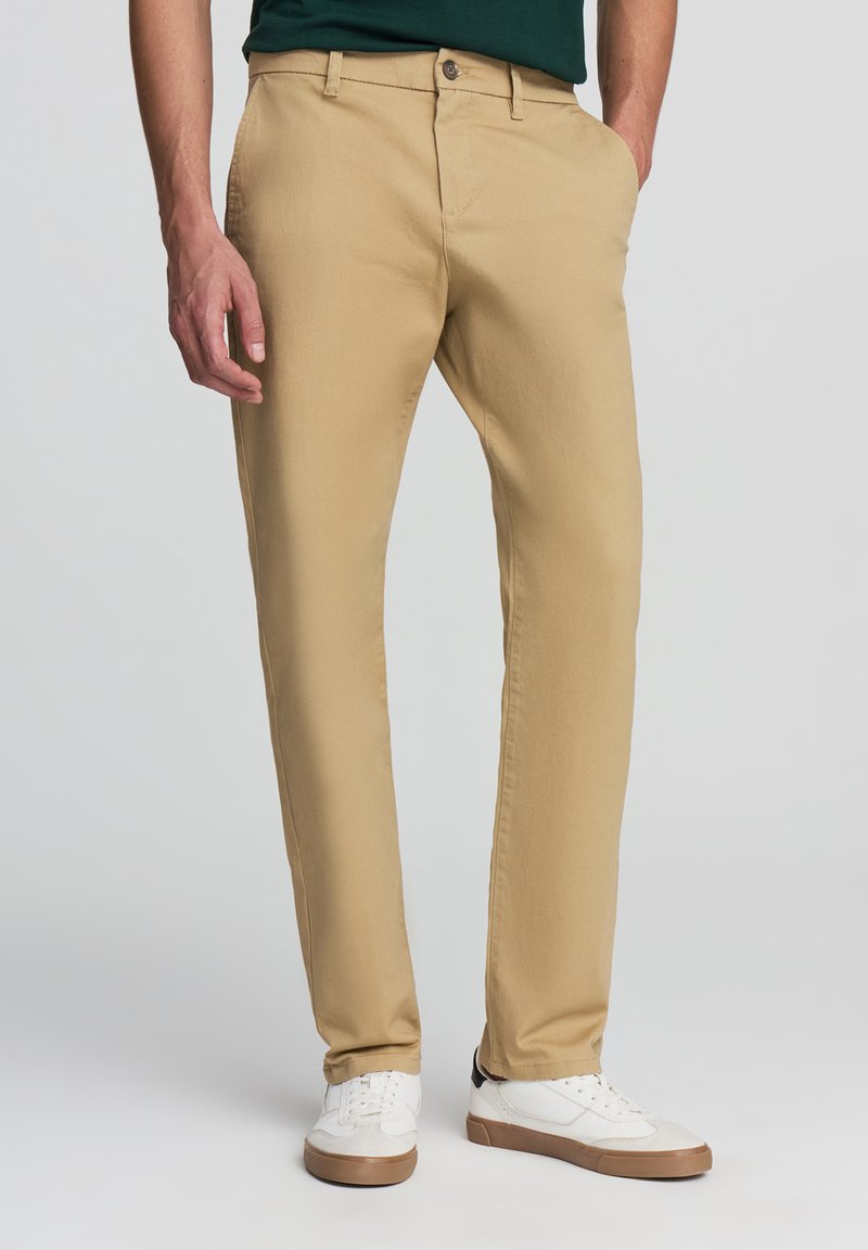 Khaki dopasowane chinosy wykonane z tkaniny bawełnianej. Posiadają klasyczny pięciopocketowy fason, zapięcie na guzik oraz gładką teksturę.