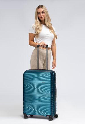 Valise rigide bleue avec des rainures diagonales, poignée noire et quatre roues. Design compact avec une finition brillante et un logo à l'avant.