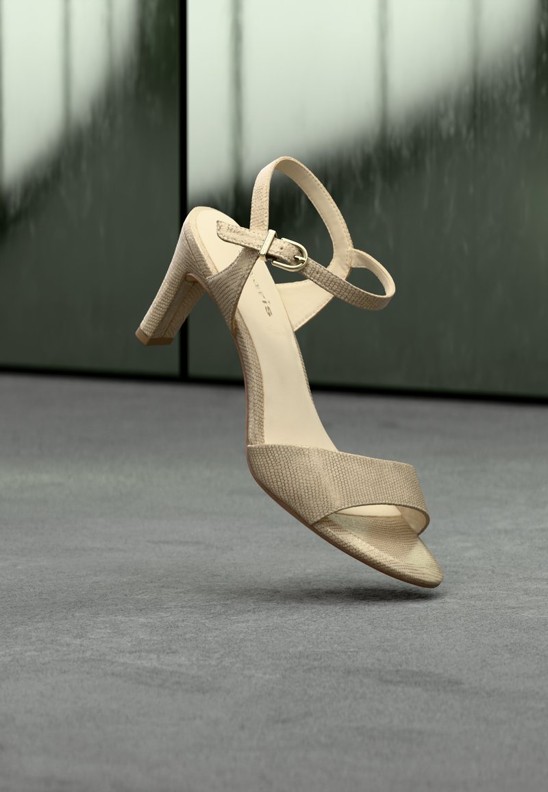Beige high-heeled sandal with ankle strap and buckle, textured pattern, shown mid-air against a blurred green and gray background.