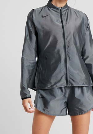 Nike Windbreaker in Dunkelgrau, mit Reißverschluss vorne, hohem Kragen und einem glatten, glänzenden Stoff. Kombiniert mit passenden Shorts, ebenfalls in Grau.