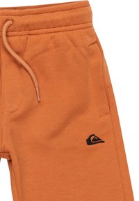 Pantalones de chándal naranjas hechos de una tela suave, con una cinturilla elástica con cordón y un pequeño detalle de logo negro en el muslo izquierdo.