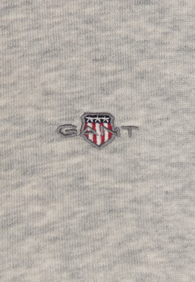 Un tissu tricoté gris arbore un petit logo brodé avec un design de bouclier, incorporant des accents rouges et bleus.