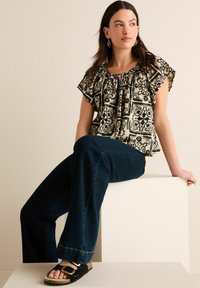 Blouse à motifs floraux crème et noir avec des manches flottantes, associée à un jean évasé bleu marine et des sandales noires avec des accents dorés.