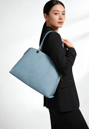 Donna in completo nero che tiene una grande borsa azzurro chiaro testurizzata con doppi manici sulla spalla, vista di profilo.