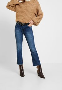 Beige villapaita, korkeavyötäröiset siniset levenevät farkut, joissa on raakareuna, ja ruskeat käärmeennahkakuosiset nilkkurit. Musta vyö korostaa vyötäröä.