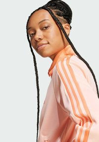 Roze atletska jakna s narančastim trostrukim trakama, visokim ovratnikom, glatkom teksturom i punim patentnim zatvaračem. Stilizirana s dugom pletenom kosom.