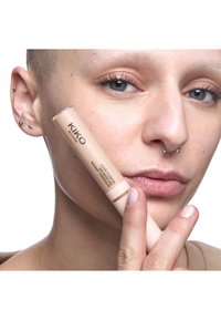Person med flere ørepiercinger og septumring holder KIKO Milano Love Fusion radiance concealer nær kinnet på hvit bakgrunn.
