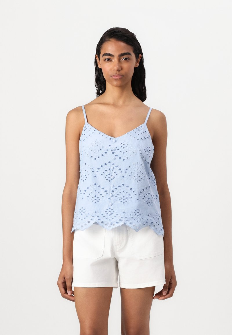 Top sem mangas azul claro com bainha recortada, apresentando padrões de ilhós bordados e alças finas ajustáveis. Usado com calções brancos.