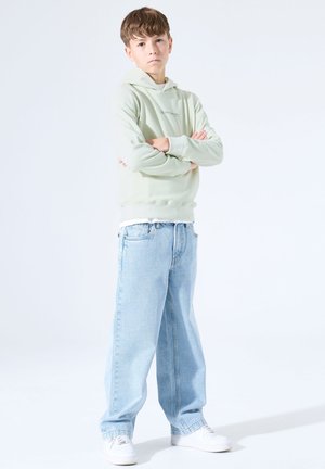 Hellgrüner Hoodie, lässig sitzende hellblaue Jeans und weiße Turnschuhe. Der Stoff wirkt weich; der Hoodie hat ein kleines Logo.