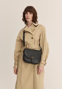 Czarna skórzana torba na ramię z zaokrąglonym klapką, złotymi akcentami i regulowanym paskiem, zestawiona z beżowym trenczem.