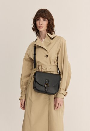 Crna kožna torba za preko ramena s zakrivljenim preklopom, zlatnim metalnim detaljima i podesivim remenom, u kombinaciji s bež trench kaputom.