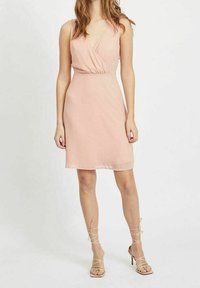 Žena v bezramenných, světle růžových zavinovacích šatech a béžových kostičkovaných vysokých podpatcích, stojící na pozadí s čistě bílou barvou.