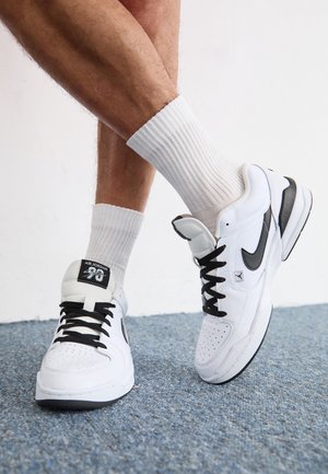 Persoon die witte en zwarte Air Jordan sneakers draagt met zwarte veters en witte geribde crew-sokken, staand op een blauw tapijt.