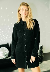 Blondýnka v černých šatech na knoflíky se zdobením stojí v moderní místnosti s diskovou koulí, která odráží světlo.