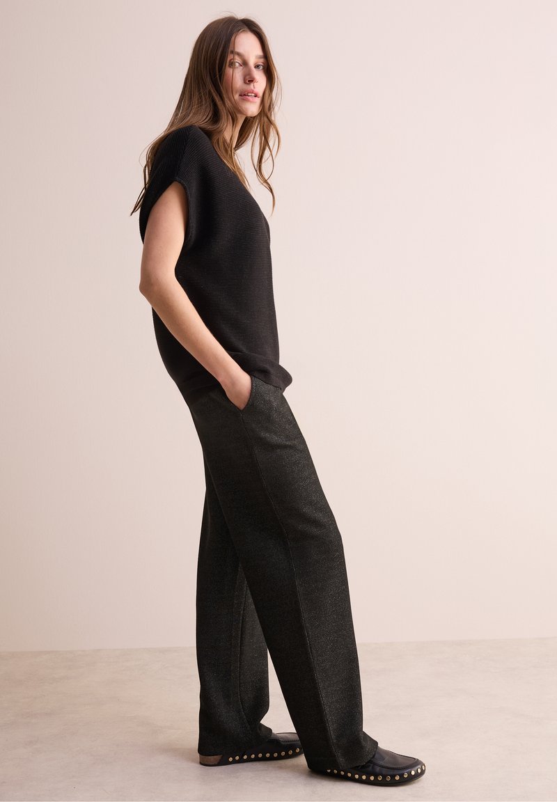 Zwarte geribbelde korte mouwen top, gecombineerd met losse, donker gestructureerde broek. Schoeisel heeft gouden studs langs de randen. Neutrale achtergrond.