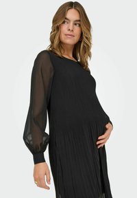 Musta midi-mekko, jossa on laskostettu pinta ja läpikuultavat, pöyhistetyt hihat. Pehmeä, virtaava kangas ja rento malli, joka on suunniteltu äitiysvaatteeksi.