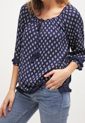 Marineblaue Bluse mit weißen geometrischen Mustern, rundem Ausschnitt, Quastenbändern und ausgefranstem Saum, kombiniert mit hellblauen Jeans.