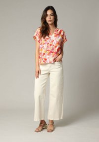 Bloes met korte mouwen in een bloemenpatroon in roze, oranje en gele tinten, gecombineerd met crème wijde broekspijpen. Sandalen met smalle straps.