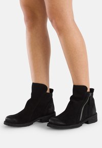 Botines de ante negros con puntera redondeada, cremallera lateral y tacón texturizado. Cuenta con un diseño de corte único en la parte superior.