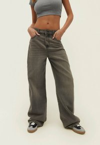 Jeans de denim gris de pierna ancha con un acabado desvanecido, que cuentan con cierre de botón, pasadores para cinturón y bolsillos laterales. Combinados con zapatillas de color contrastante.