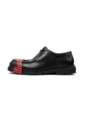 Zapato de cuero negro con punta redondeada con un tapón de goma roja. El diseño incluye cordones y una suela negra gruesa y texturizada.