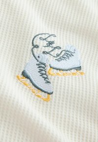 Bordado de patín de hielo blanco con cuchillas amarillas y cordones grises sobre una tela texturada color crema.