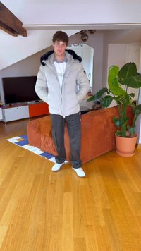 Padded grå jakke med svart pelskant, hvit T-skjorte, mørke bukser og hvite joggesko, stående i et rom med tregulv og planter.