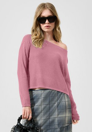Vrouw met zwarte cat-eye zonnebril, een roze off-shoulder ribgebreide trui, een grijze geruite rok, die een zwarte gerimpelde handtas vasthoudt.