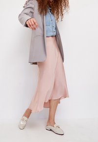 Helle graue Blazer über einem Jeanshemd, kombiniert mit einem schwingenden rosa Midirock und cremefarbenen Loafers mit goldener Akzentuierung. Glatter Stoff und taillierte Passform.